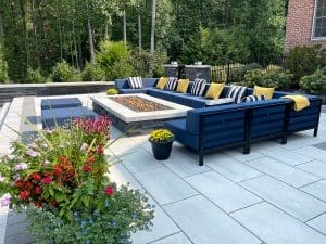 Outdoor patio with modern furniture. A blue sectional sofa with yellow and striped pillows surrounds a rectangular fire pit. Potted plants with colorful flowers are in the foreground, and greenery and trees are in the background. Stone tiles cover the patio floor.