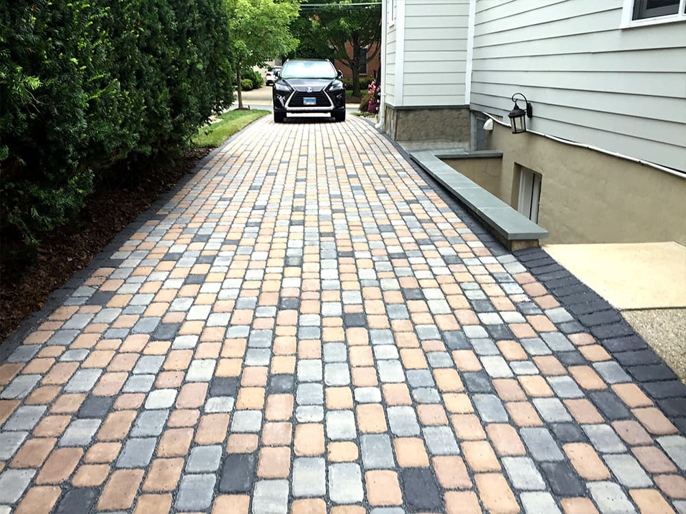 A driveway with a patterned stone pathway leads to a parked black car in front of a house. The house has light-colored siding and a side entrance next to the driveway. Green bushes border the left side of the driveway.