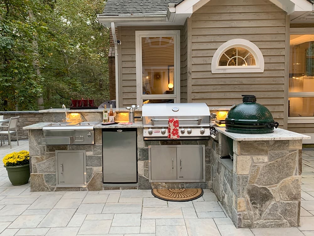 A luxurious outdoor kitchen features a stainless steel grill, a green ceramic grill, a mini-fridge, and storage cabinets set in a stone counter. The kitchen is decorated with potted yellow flowers and illuminated string lights. Trees and a house are in the background.