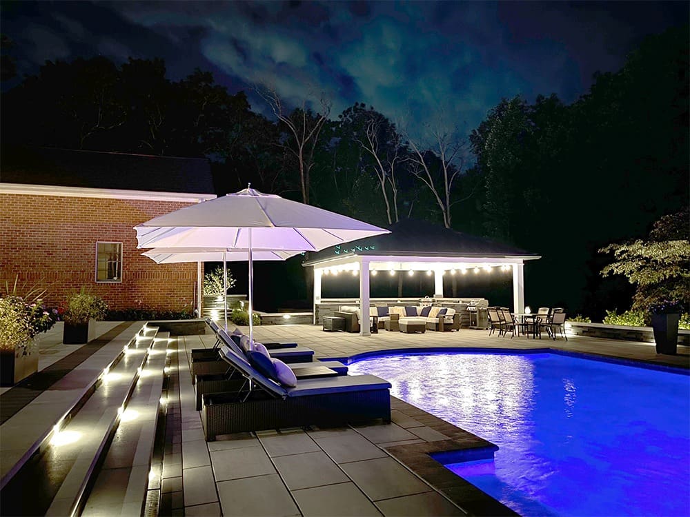 A luxurious backyard pool area at night, featuring a glowing blue pool surrounded by modern lounge chairs with a large white umbrella. A brightly lit gazebo with seated dining area is in the background, with trees and a partially visible house adding to the serene ambiance.