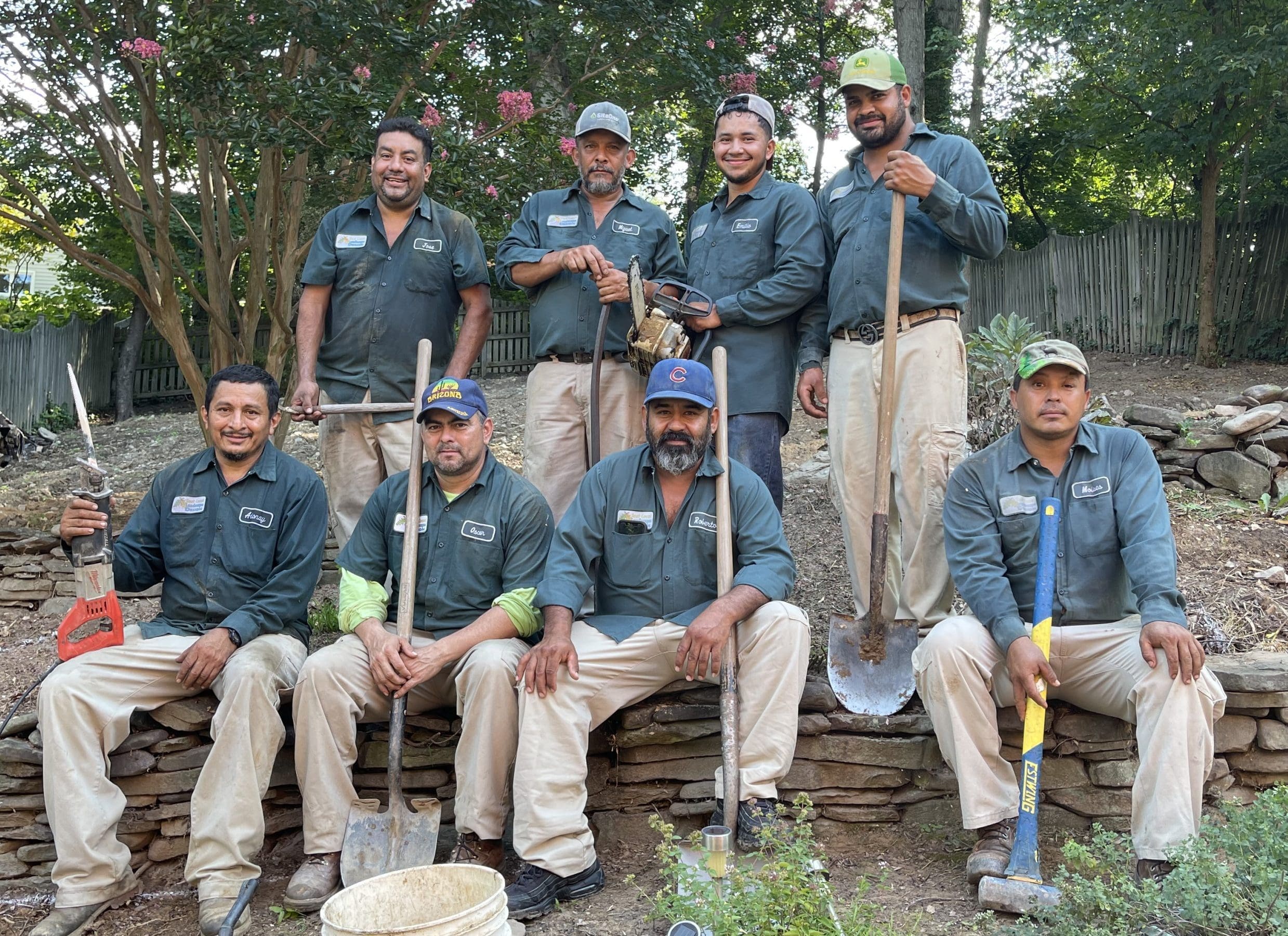 A group of eight landscapers poses together outdoors. Seven are standing in the back row, each holding a shovel or a tool, while one kneels in the front holding a rake. They're wearing matching work uniforms consisting of green shirts and beige pants. A wooded area is in the background.