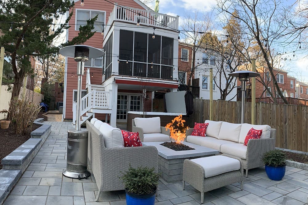 A backyard with patio furniture arranged around a rectangular fire pit. The patio is paved with stone tiles and includes two outdoor heaters. A multi-story house with a balcony and a screened porch is in the background. There are bare trees and potted plants.