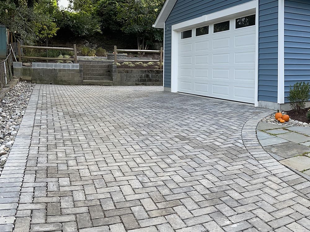 A clean and neatly paved driveway made with rectangular interlocking bricks leads up to a blue house with a white garage door. Along the driveway, there is a border of small, white rocks. The garden has a few plants and bushes, with a retaining wall at the far end.