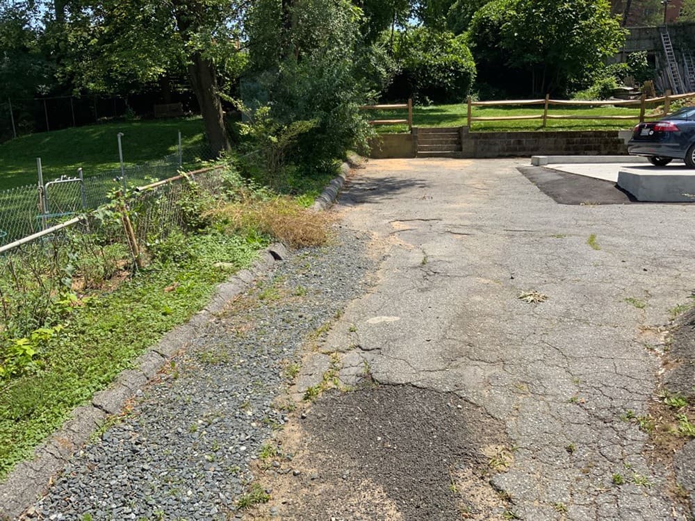 Driveway BEFORE Horizontal 1000 x 750 An outdoor, cracked asphalt driveway transitions to a gravel path on the left. The area is bordered by a chain-link fence and greenery. Trees and a wooden fence are in the background, with a grassy area slightly elevated beyond stone steps. A parked car is on the right.