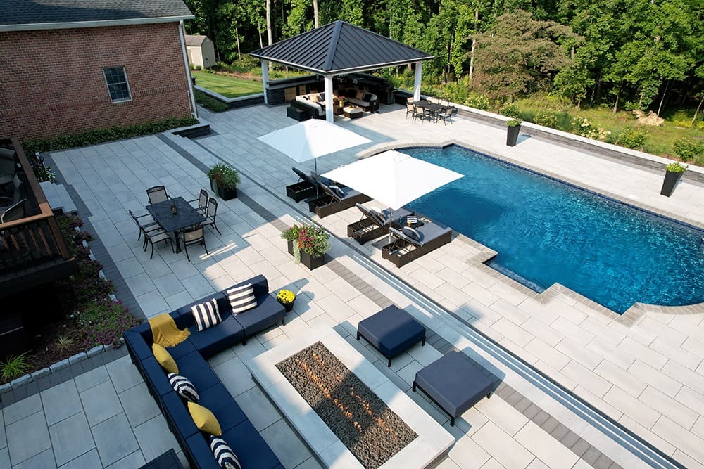 A spacious backyard features a modern pool surrounded by large, light-colored tiles. There are white umbrellas, black cushioned outdoor furniture, and a dining table. A poolside pavilion with additional seating is in the background, near trees and greenery.