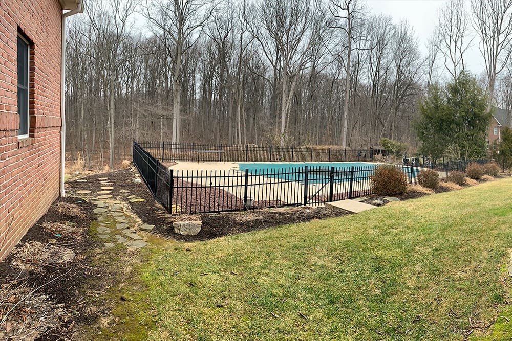 A partially empty backyard with a rectangular in-ground pool surrounded by a black metal fence. The pool area is adjacent to a wooded area of bare trees. The house, made of red brick, is visible on the left side. The lawn in the foreground is bordered by mulch and stepping stones.