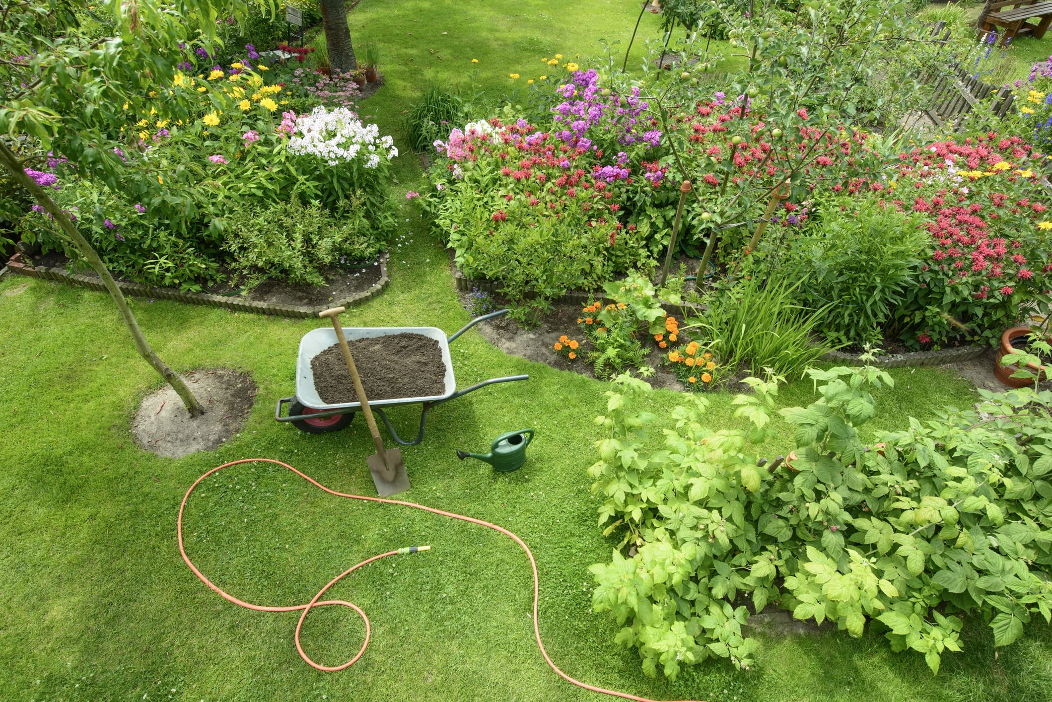 A lush garden with various flowers and plants, including a bed of purple, pink, red, and yellow blooms. A wheelbarrow filled with soil, a shovel, a green watering can, and an orange garden hose are placed on the neatly mowed lawn.