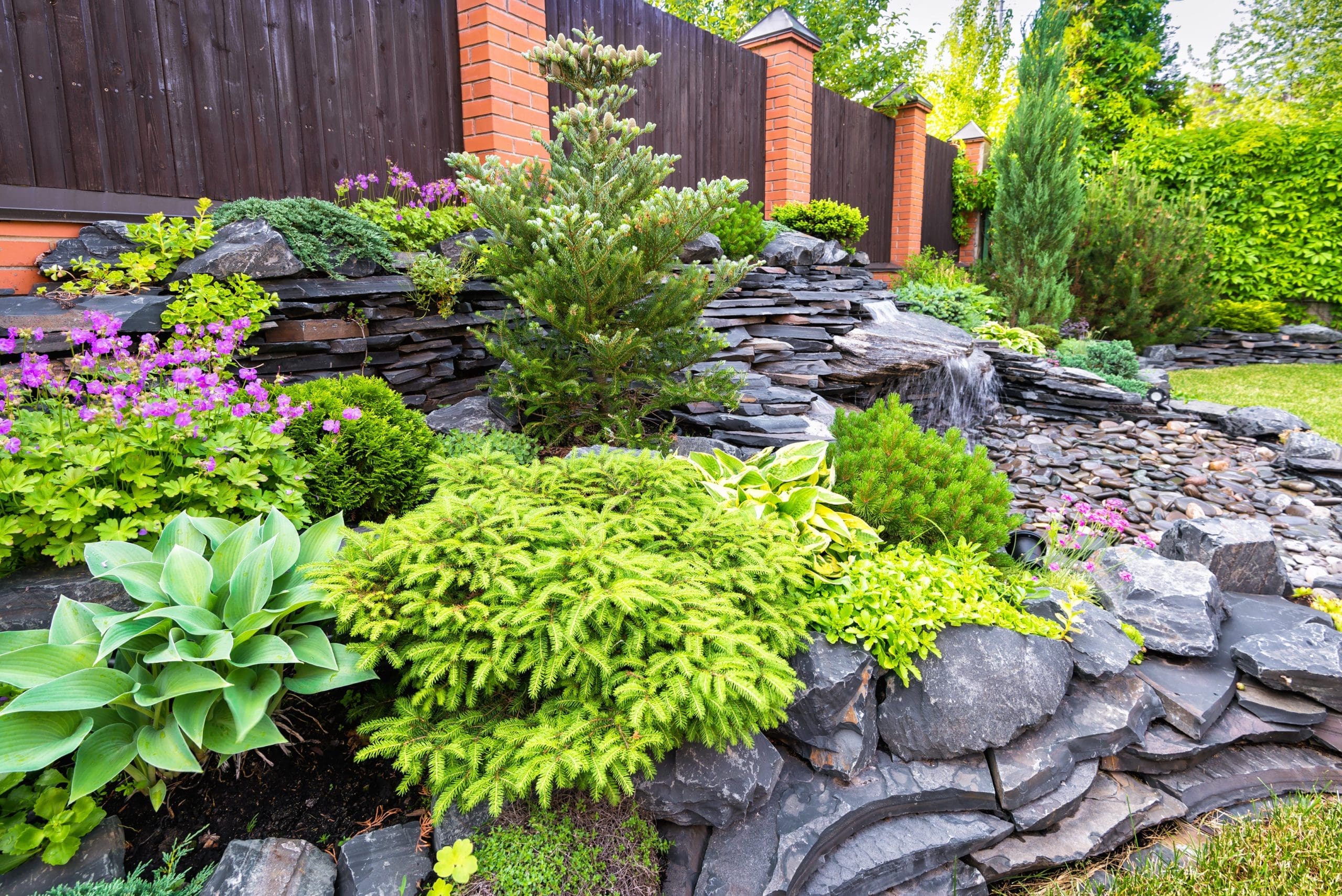 A well-maintained garden with various lush green plants, including hostas and conifers, surrounded by layered stone landscaping. In the background, there is a wooden fence with brick posts.