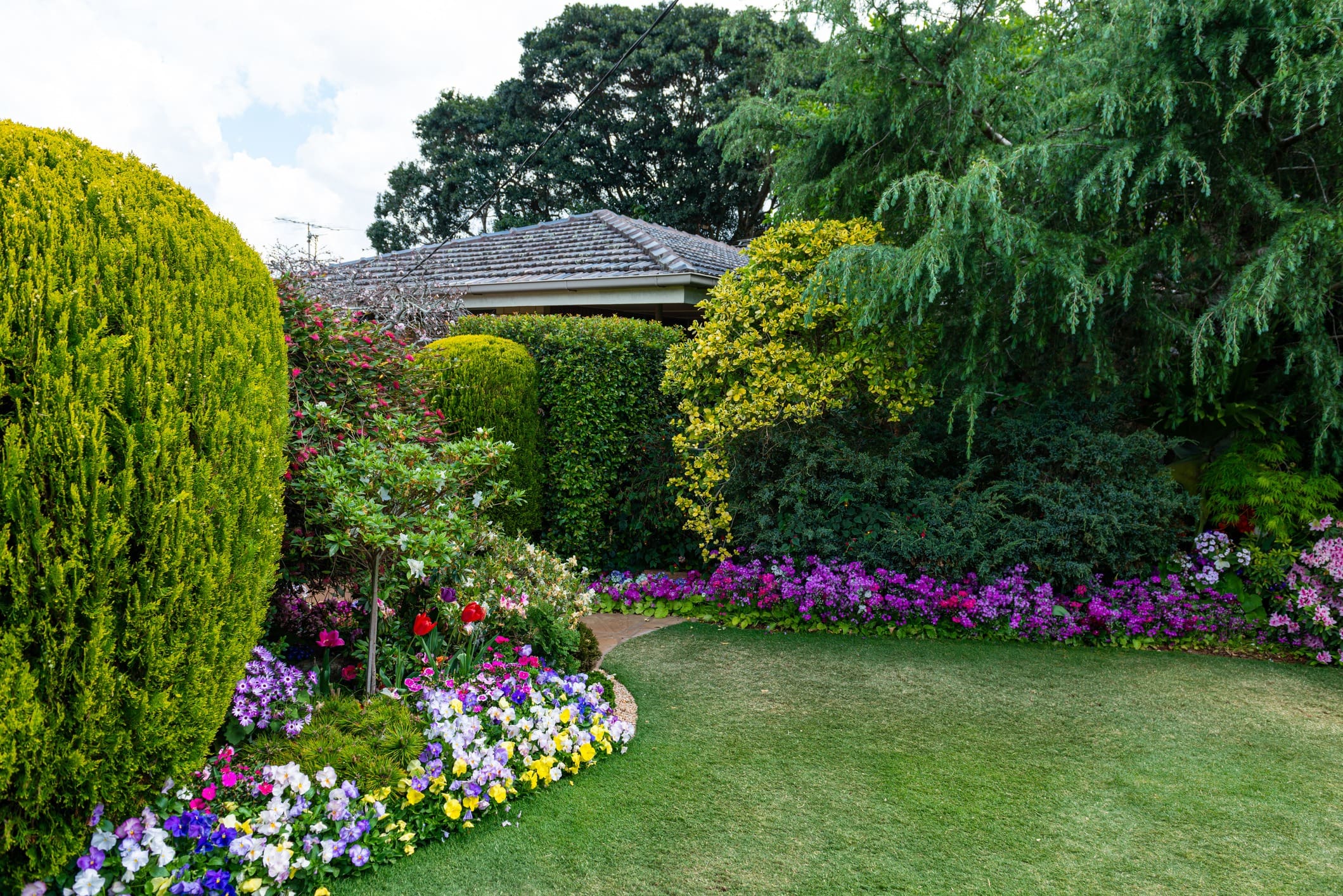 A lush garden with a variety of colorful flowers, including purple, yellow, and white blooms, surrounds a house with a tiled roof partly obscured by trees and bushes. The well-manicured lawn leads up to the vibrant flower beds and dense greenery.