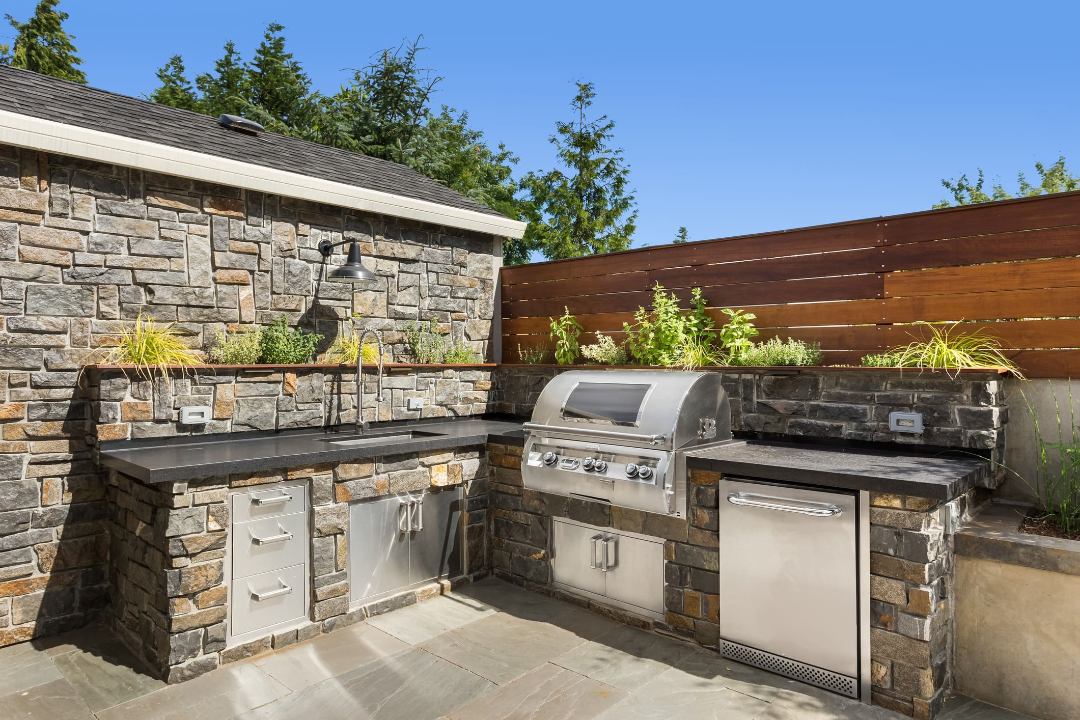 Outdoor kitchen featuring a stainless steel grill, sink, and storage set against a stone counter and stone wall backdrop. The kitchen is adorned with green plants and bordered by a wooden fence, with trees and clear blue sky in the background.