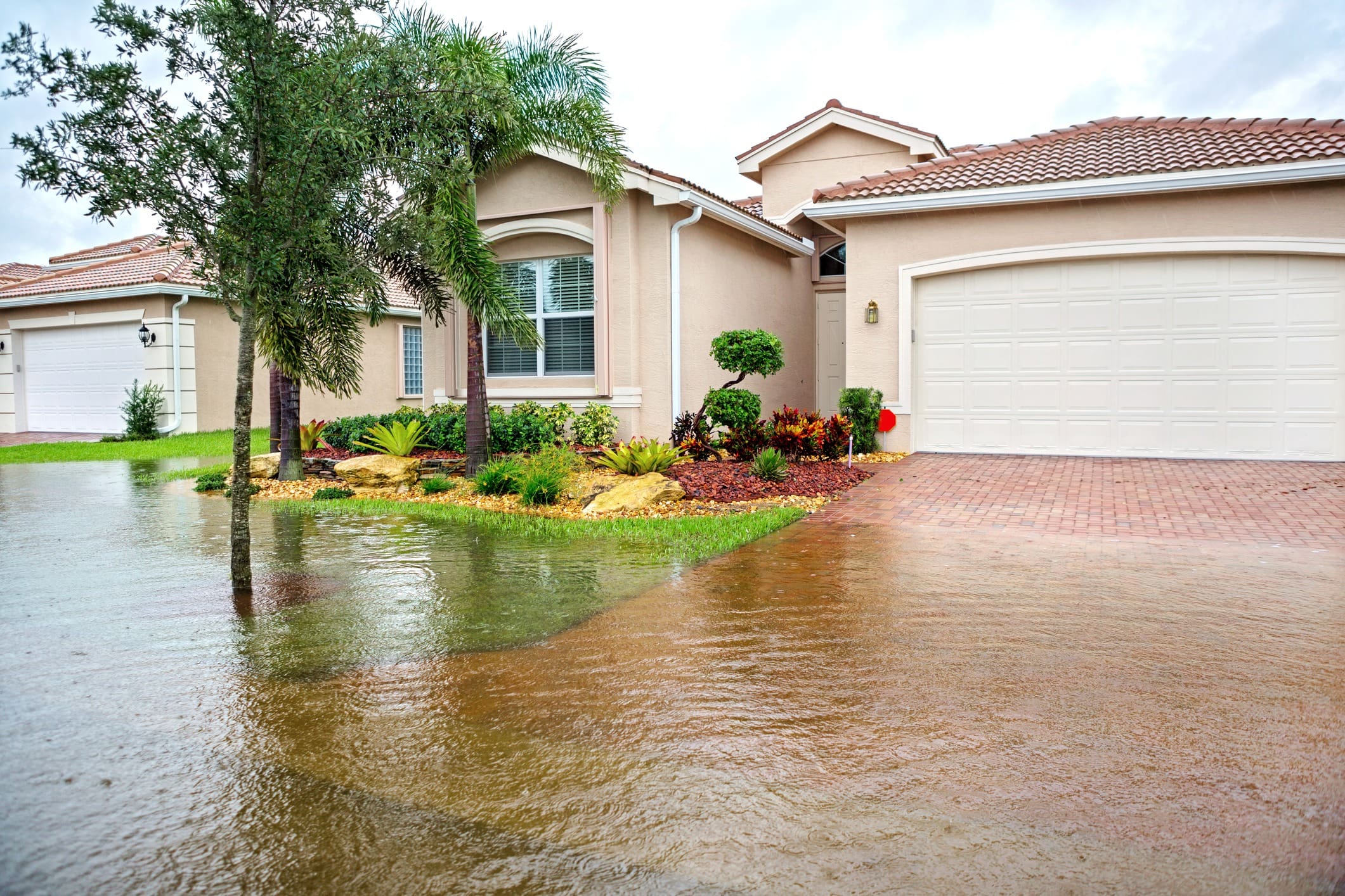 A single-story house with a red-tiled roof is seen with floodwaters covering the driveway and front yard. The garage door is closed, and there are palm trees and shrubs in the landscaping. The sky appears overcast, suggesting recent or ongoing rain.