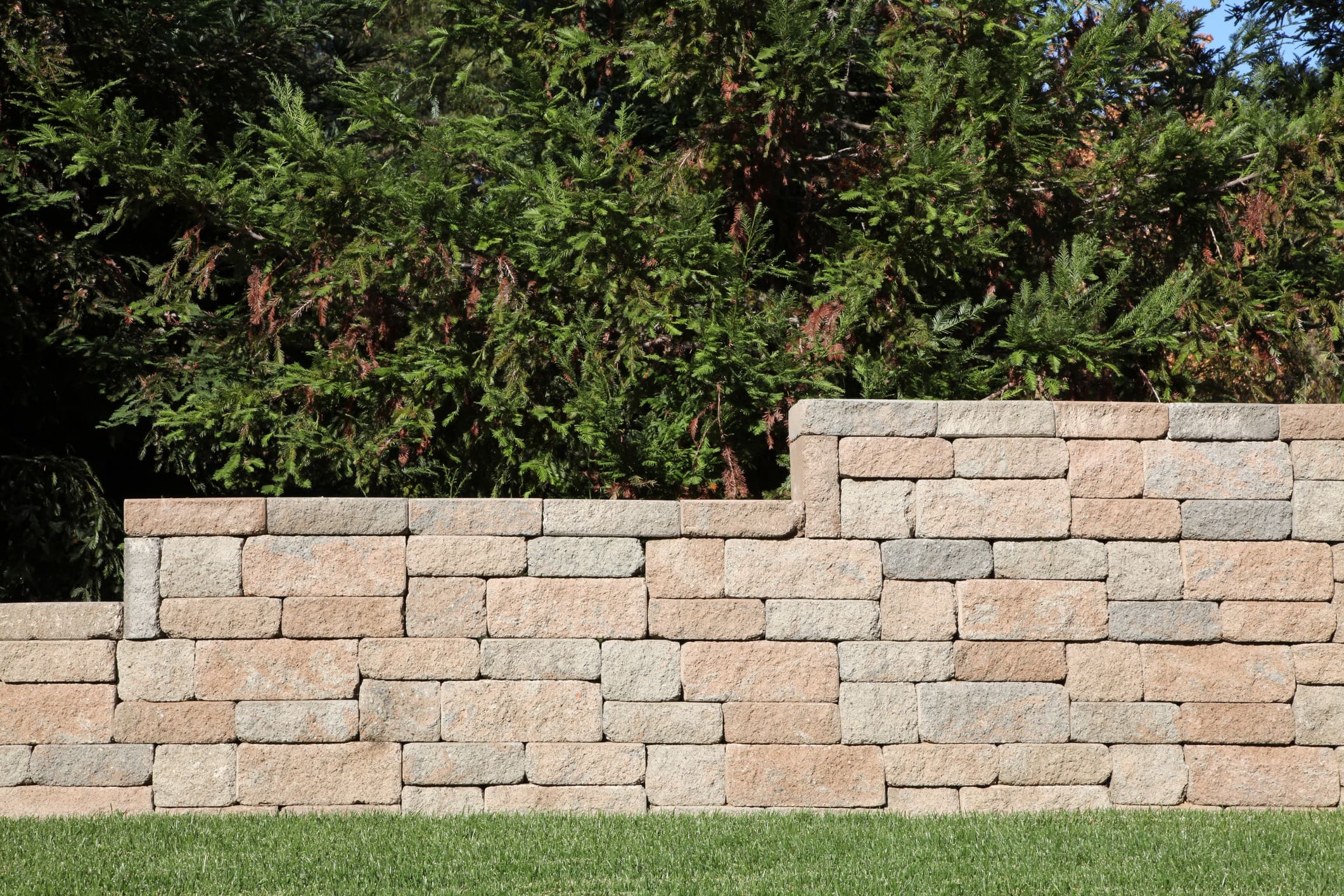 A sturdy stone retaining wall with varying heights stands in front of lush green foliage. The neatly stacked rectangular stones contrast with the vibrant greenery, while a well-manicured lawn stretches out in the foreground.