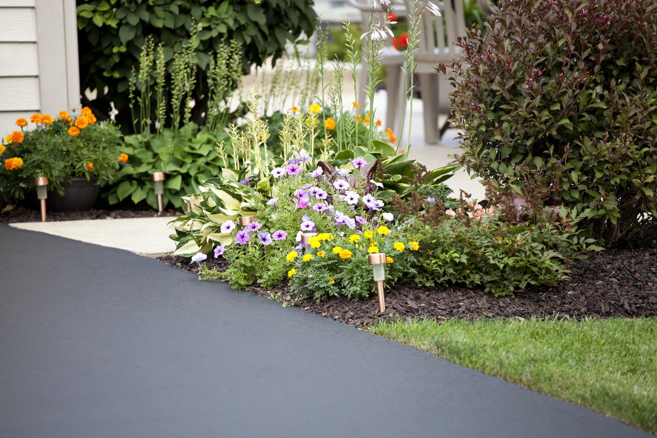 A well-manicured garden featuring marigolds, petunias, and other colorful flowers borders a freshly paved driveway. Small pathway lights are spaced along the flowerbed's edge. In the background, there is lush greenery and outdoor furniture.