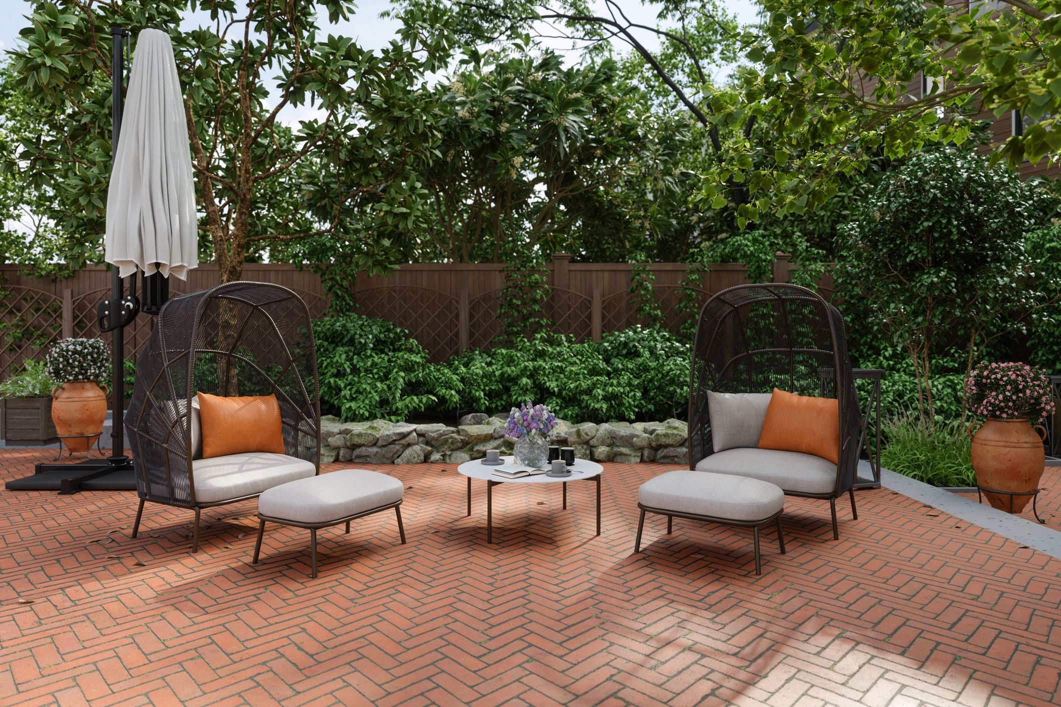 A cozy outdoor patio with brick flooring features two cushioned lounge chairs with orange throw pillows and matching ottomans. A small round table with flowers and mugs sits between them. An umbrella and lush greenery surround the scene, creating a tranquil atmosphere.