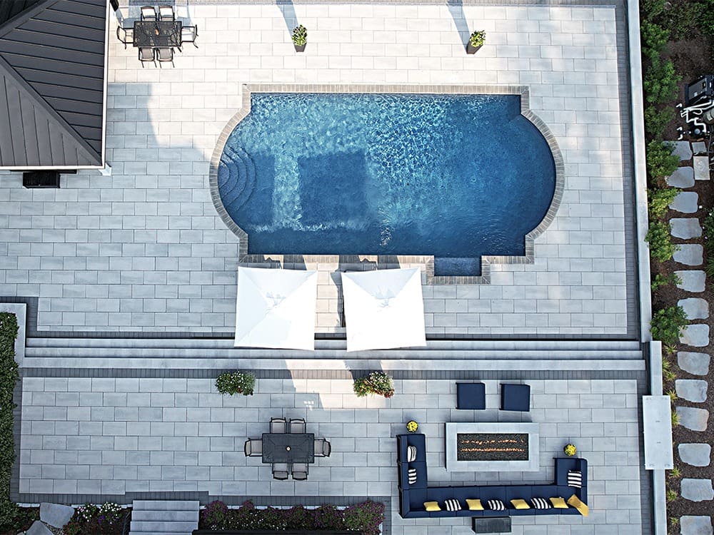 Aerial view of a backyard featuring a rectangular swimming pool with rounded corners, surrounded by a stone patio. Two white umbrellas are set up on the pool's left side. Adjacent, there is patio furniture including a dining set and a lounge area with a fire pit.