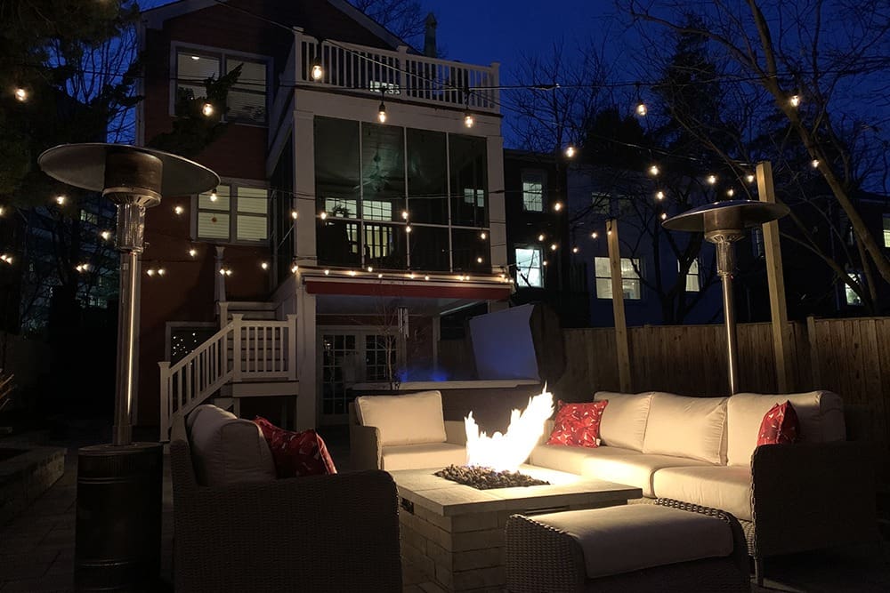A cozy backyard patio at night featuring a fire pit surrounded by sectional seating and string lights hanging overhead. A two-story house with lit windows is in the background, adding to the inviting ambiance of the outdoor space.