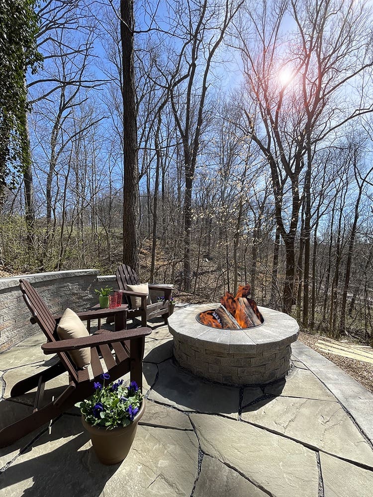 A cozy outdoor patio with two wooden chairs and a fire pit. The patio is surrounded by a tranquil wooded area with bare trees, under a clear blue sky. There is a potted plant with purple flowers next to the chairs, and sunlight filters through the trees.