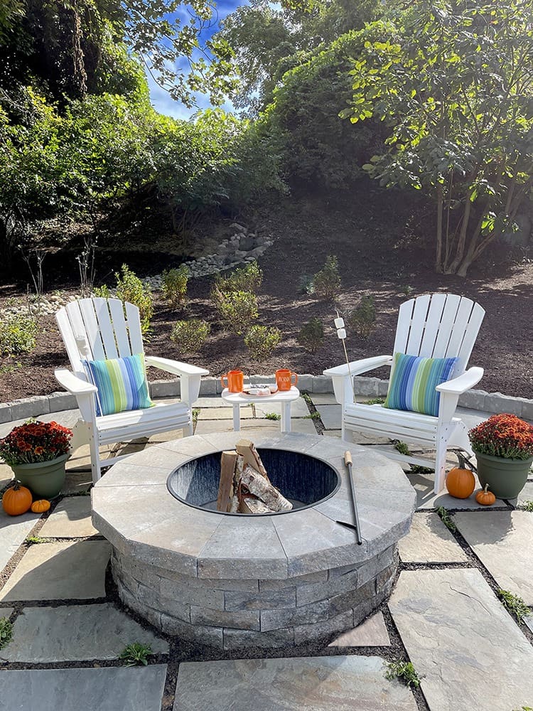 A serene backyard setting featuring two white Adirondack chairs with striped pillows placed around a stone fire pit. Two mugs sit on side tables near the chairs, surrounded by potted plants with mums and small pumpkins. Lush greenery and trees are visible in the background.