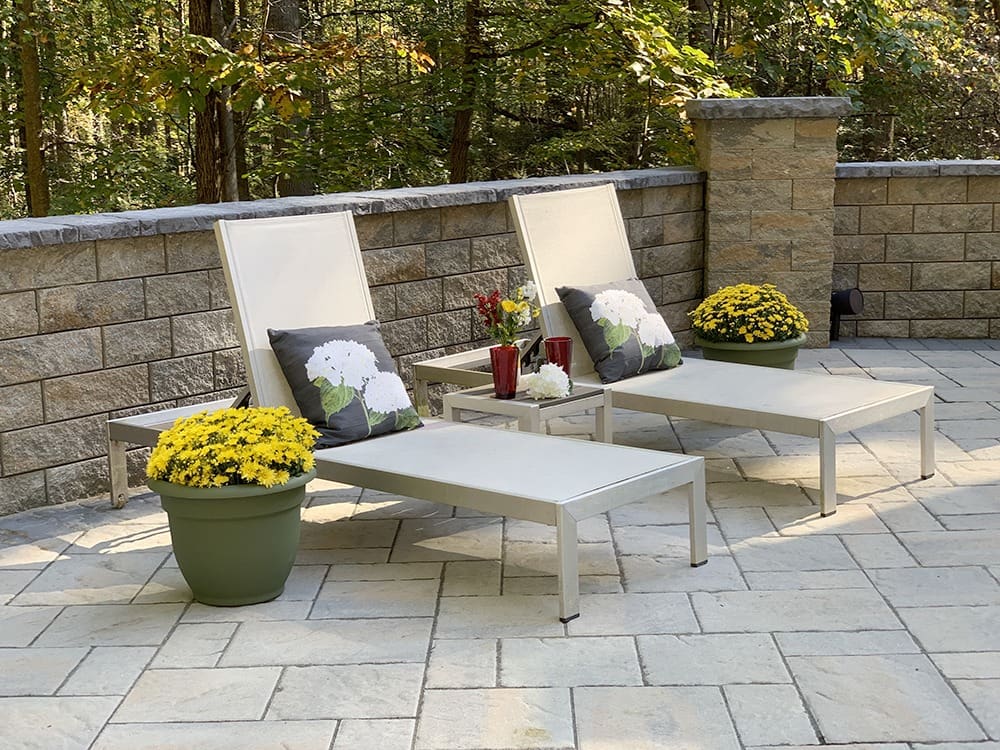 Two reclining lounge chairs with floral pillows are arranged side by side on a tiled patio. Bright yellow potted chrysanthemums are positioned at the head of each chair. The patio is bordered by a stone wall with trees visible in the background.