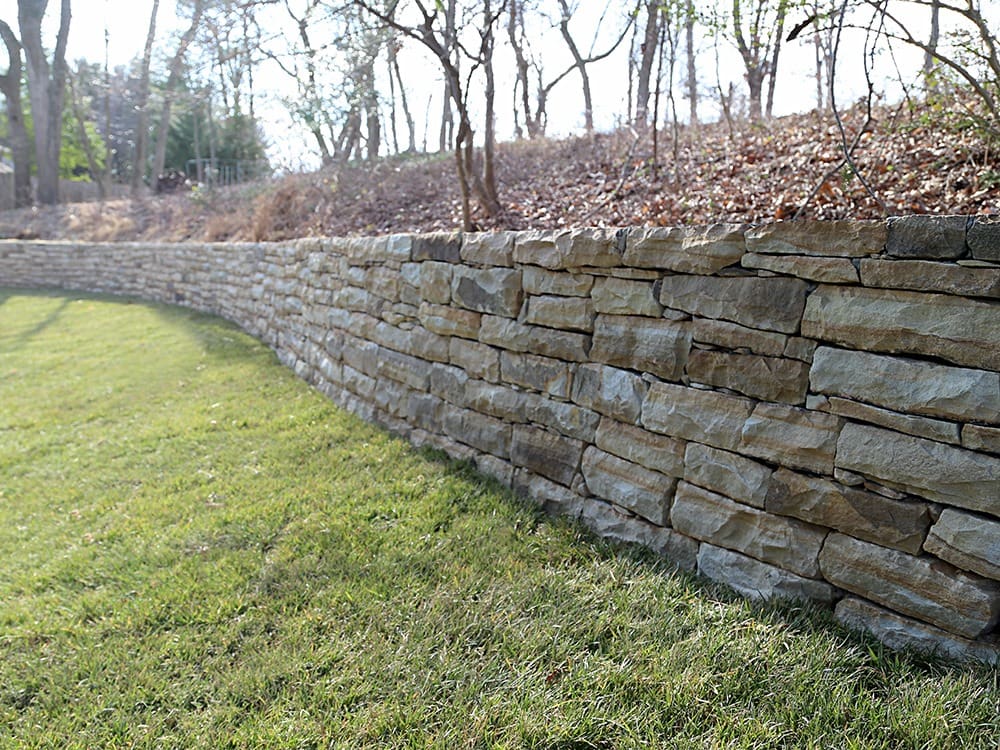 A stone retaining wall curves along a grassy slope, with bare trees and slightly wooded area in the background. The wall is built from neatly stacked, rectangular stones.