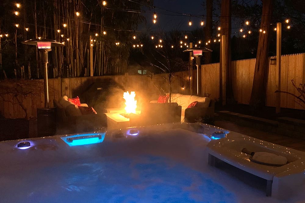 A backyard scene at night featuring a bubbling hot tub in the foreground, glowing with blue light. In the background, there are two chairs surrounding a fire pit. String lights hang above, illuminating the cozy area. Two tall heaters are placed next to the chairs.