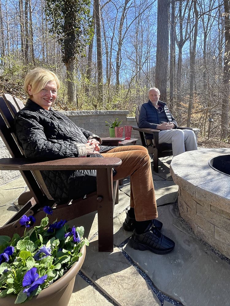 Two elderly people sitting on wooden chairs in an outdoor setting. The woman is wearing a black quilted jacket and brown pants, and the man is wearing a gray jacket and light pants. There is a round fire pit in front of them and a potted plant with purple flowers nearby.