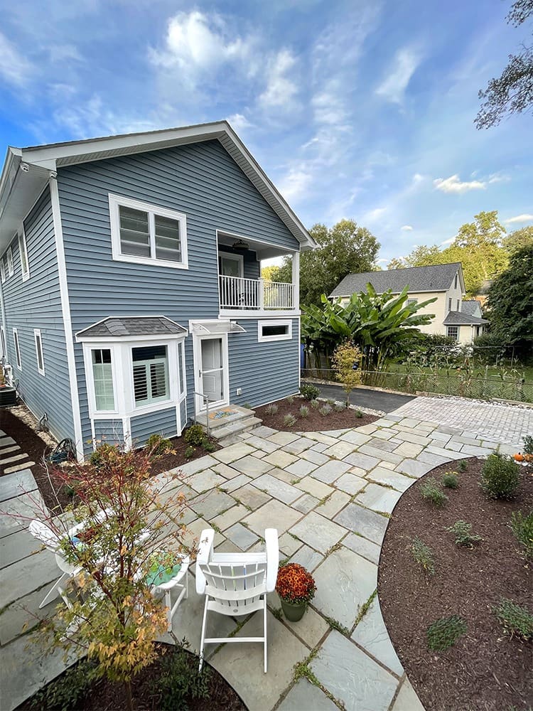 Two-story blue house with white trim viewed from the backyard. The yard features a stone patio with white chairs and a small tree surrounded by mulch. Other plants and a neighboring house are visible in the background, all under a cloudy sky.
