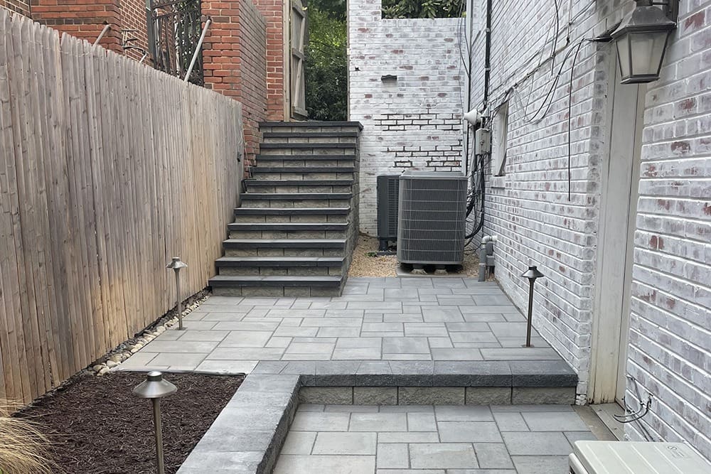 A narrow outdoor area with gray paving stones, bordered by brick walls on the left and right. A set of stairs leads to a higher level at the back, bordered by a wooden fence on one side. Two HVAC units sit near the brick wall on the right. Pathway lights are along the steps.