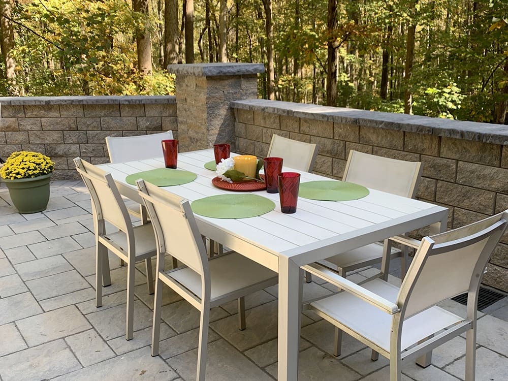 An outdoor patio with a white dining table set for six. The table features green placemats, red glassware, and a decorative centerpiece with candles. A potted plant with yellow flowers is in the corner. The area is surrounded by trees and a stone wall.