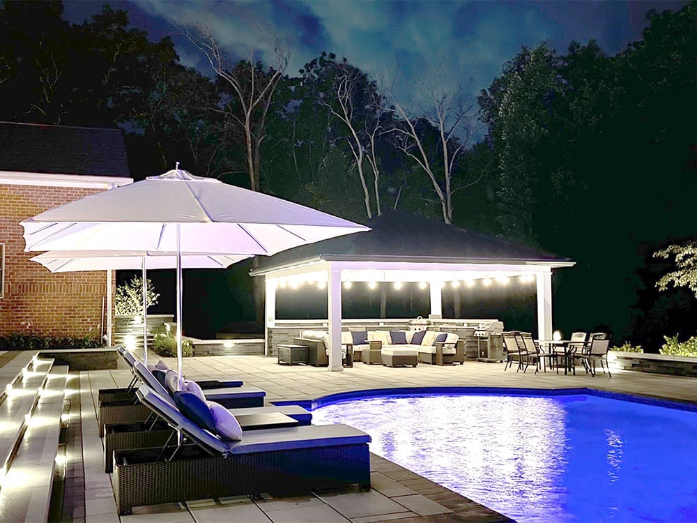 A backyard illuminated at night features a glowing pool surrounded by lounge chairs with cushions, one shaded by a large umbrella. A light-strung pergola sits in the background, sheltering a dining table and a seating area with couches. Trees form a dark backdrop.