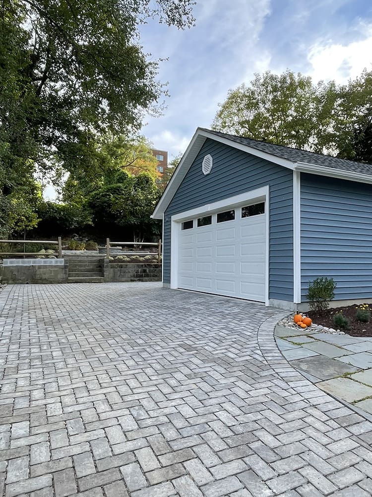 A blue detached garage with a white door stands beside a paved driveway. The yard is landscaped with trees and a small garden to the right near the building. A few pumpkins are placed near the garden, suggesting an autumn season. The sky above is partly cloudy.
