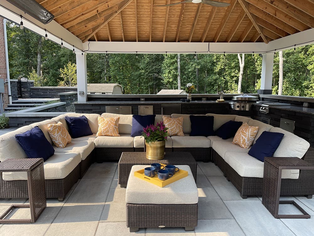 A cozy outdoor patio features a large, L-shaped wicker sectional sofa with cream cushions and blue and yellow pillows. A small ottoman with a brass tray holding cups and flowers sits in the center. The space is under a wooden pergola with a built-in kitchen.