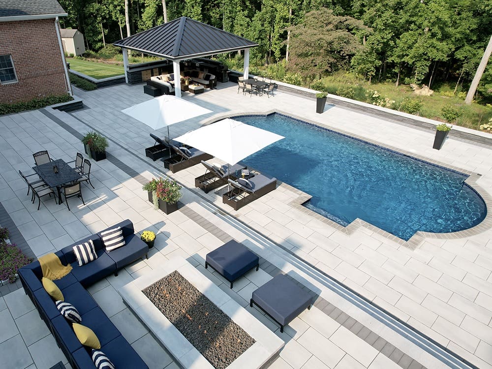 Aerial view of a modern backyard featuring a swimming pool with light gray tiles, white umbrellas, and poolside loungers. A shaded pavilion with seating is in the background. A brick house and an outdoor seating area with blue furniture and cushions are also visible.