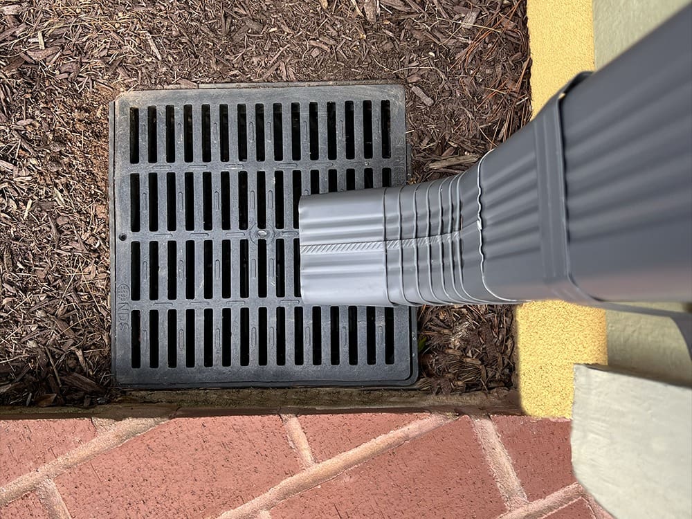 A close-up view of a gray downspout directing water into a black metal drainage grate, which is embedded in the ground surrounded by brown mulch and bordered by a yellow building wall and red brick pavement.