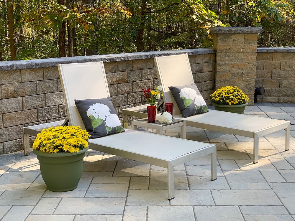 Two white lounge chairs with floral cushions are positioned on a stone patio next to a low brick wall. Each chair is flanked by large pots of yellow chrysanthemums, and small red potted plants sit on the side tables beside the chairs. Trees are visible in the background.