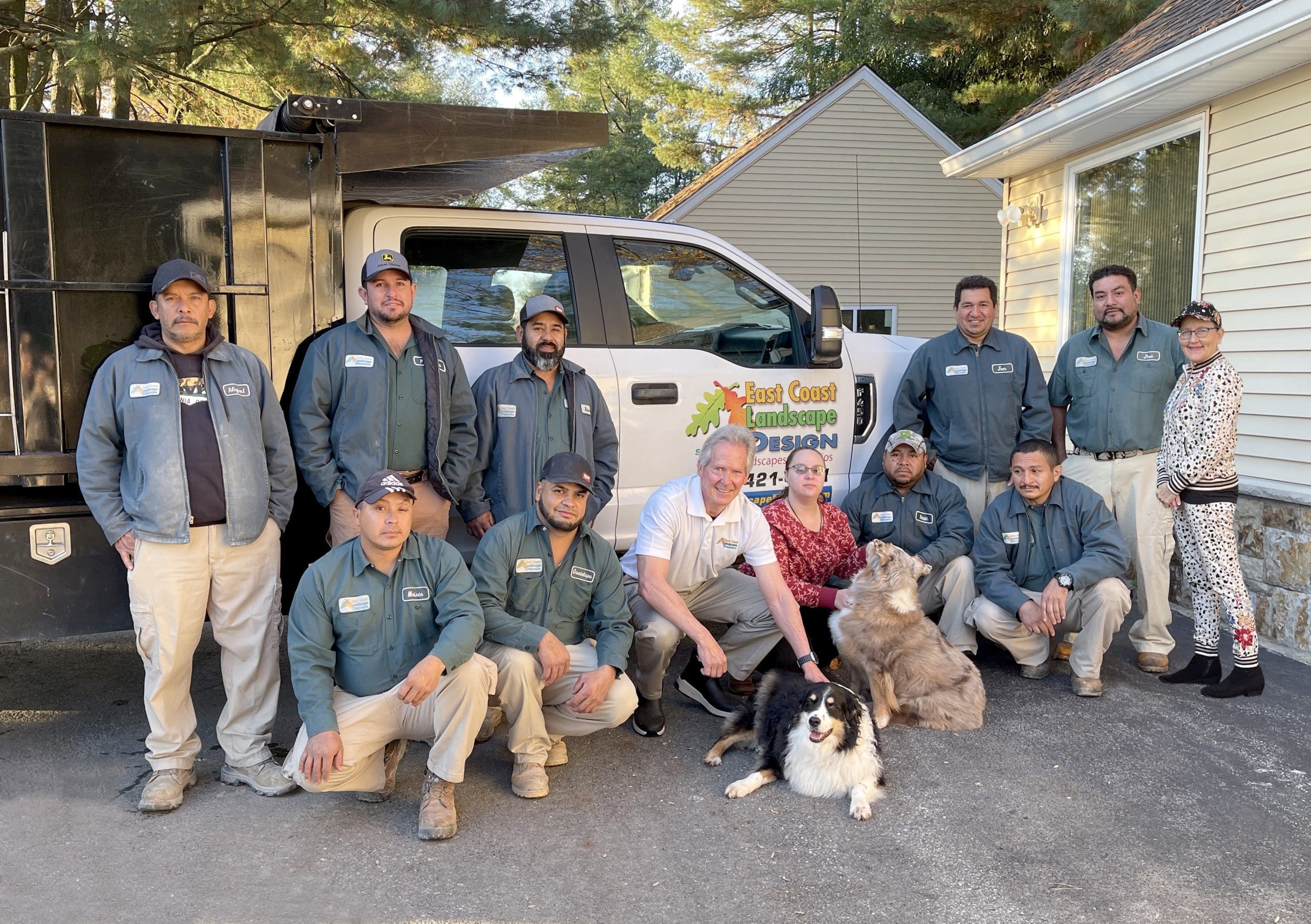 A team of eleven people and two dogs is gathered in front of a white truck with the logo "East Coast Landscape Design." The team, dressed in gray jackets, poses for a group photo on a driveway, with two people crouching and the rest standing.