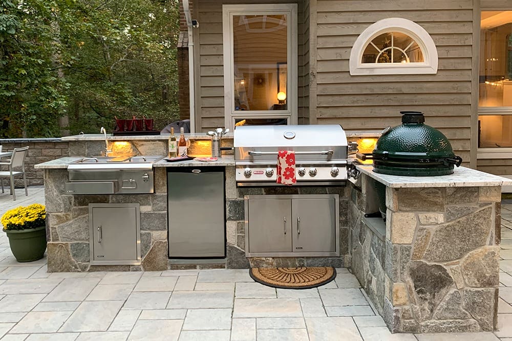 An outdoor kitchen with stone countertops and a built-in grill area. The space includes a stainless steel grill, green ceramic smoker, sink, small refrigerator, and storage cabinets. The area is surrounded by stone tiles and adjacent to a house with a circular window.