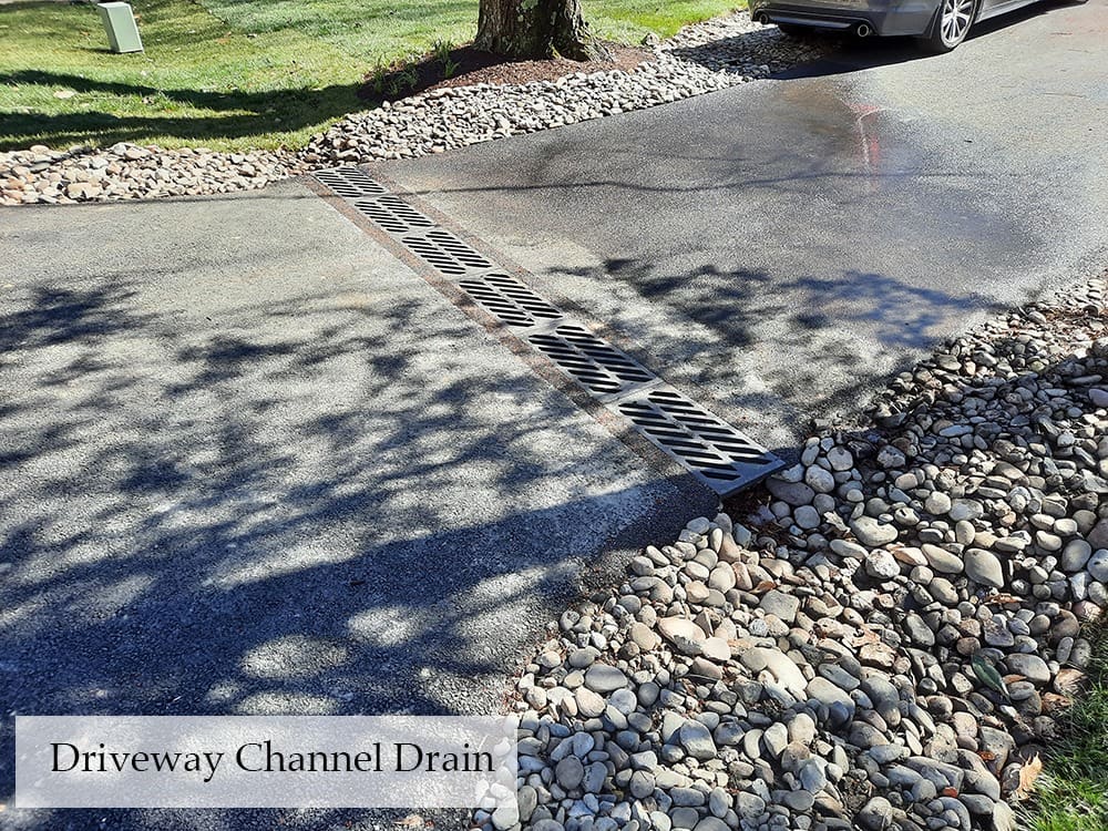 A driveway with a channel drain across its width, designed to catch water runoff. The drain has a metal grate and the driveway is bordered by a bed of small rocks. There's a car parked to the right, with a large tree in the background casting shadows on the ground. Text reads "Driveway Channel Drain".