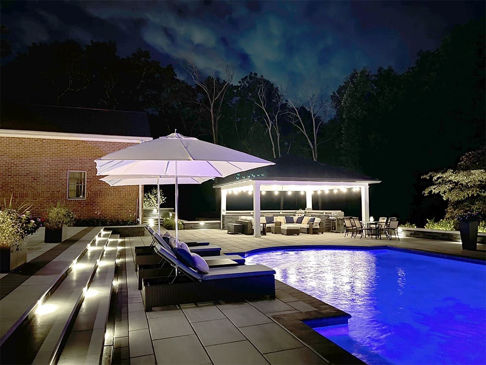 Nighttime view of a luxurious outdoor pool area, featuring illuminated pool water, lounge chairs under a large white umbrella, steps with LED lights, and a covered seating area with string lights in the background. Trees and a brick building are in the background.