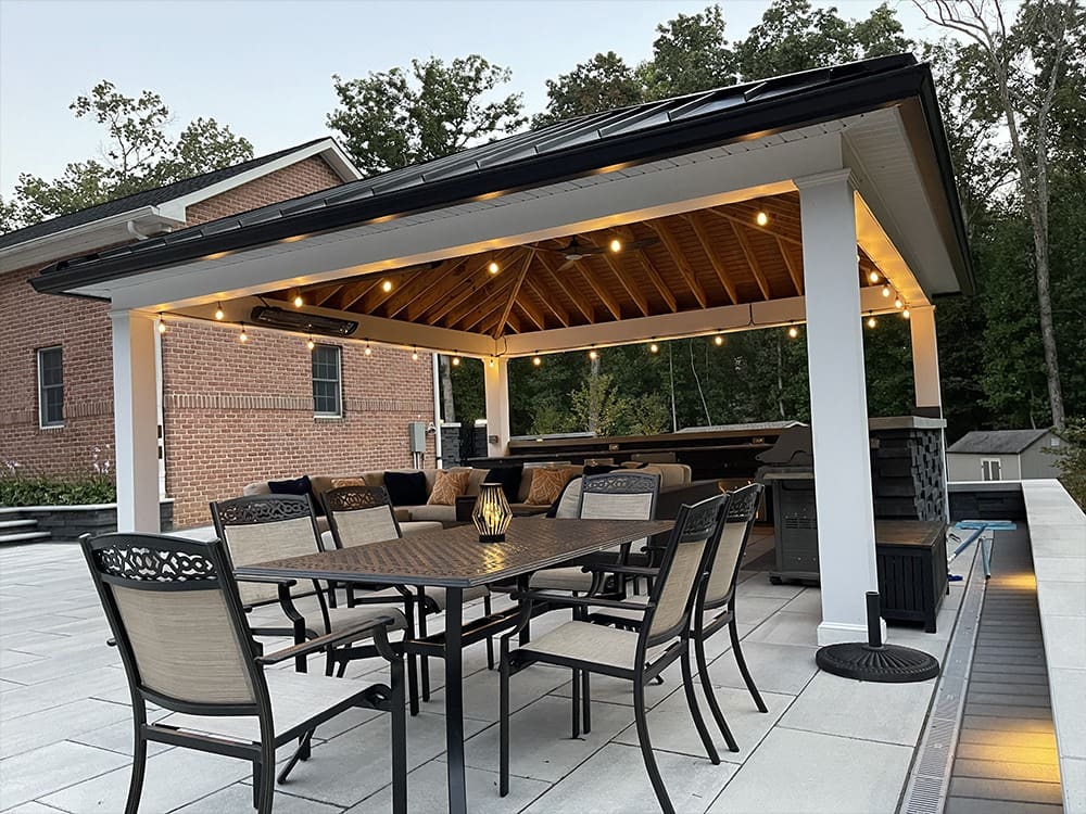 A spacious outdoor patio features a wooden pavilion with string lights hanging from the roof. Underneath, a table with eight chairs is set up for dining. The background includes a brick house, a hot tub, and dense greenery in the distance.