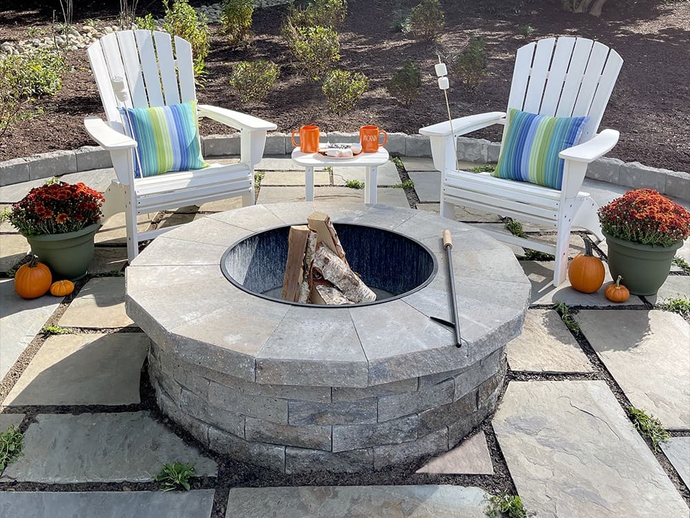 A cozy outdoor fire pit area with two white Adirondack chairs, each adorned with a colorful striped pillow. The fire pit is surrounded by stone tiles, and there's a roasting stick, potted plants, pumpkins, and two mugs placed on the chairs' armrests.