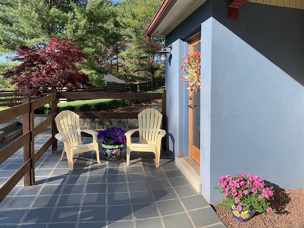 A cozy patio with two yellow Adirondack chairs flanking a colorful flower pot sits outside a blue house. The door is adorned with a festive wreath. To the right, more vibrant potted flowers are in bloom under clear skies. Lush greenery surrounds the area.