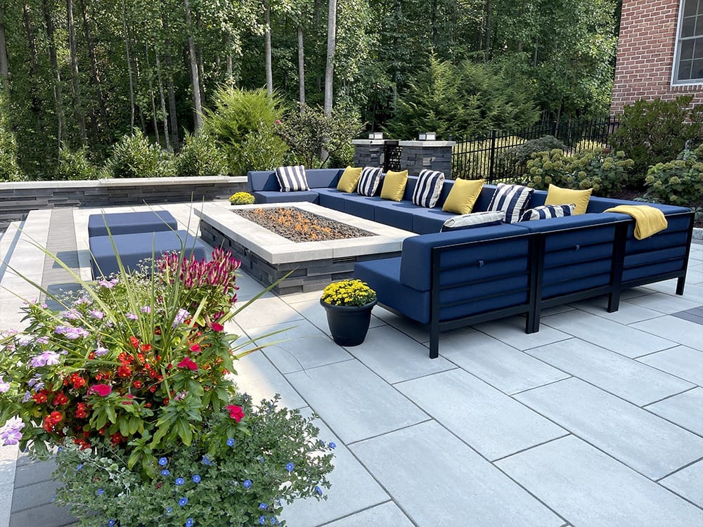 A spacious outdoor patio featuring a modern rectangular fire pit surrounded by a blue cushioned sectional sofa adorned with striped and yellow pillows. Lush foliage and a variety of potted plants enhance the tranquil setting.