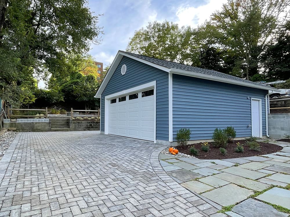 A blue detached garage with white trim featuring two large white garage doors. The surrounding area includes a newly laid brick driveway, a small landscaped garden with shrubs and pumpkins, and a terrace with retaining walls and railings at different levels. Trees stand in the background.