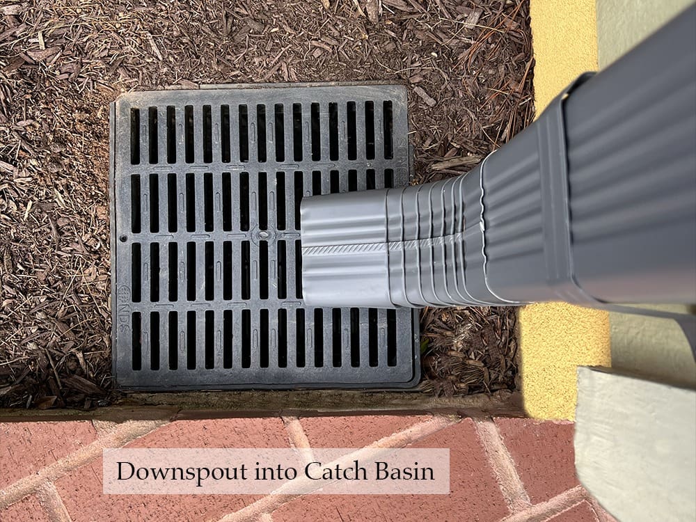 A downspout channels rainwater into a catch basin, which is a black rectangular drainage grate set into the ground, surrounded by soil on one side and paved bricks on the other. A small text label reads "Downspout into Catch Basin.